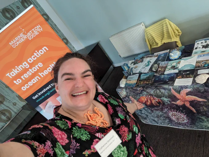 Selfie of Catherine wearing another marine themed dress and octopus necklace with a marine conservation banner behind her and a stall of leaflets with a table cloth of marine creatures