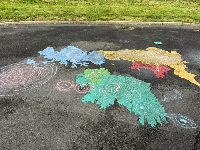 A photo of a playground with a multicoloured map of the uk printed onto the playground. Chalk rock art motifs, drawn by the pupils can be seen on the ground and across the map.