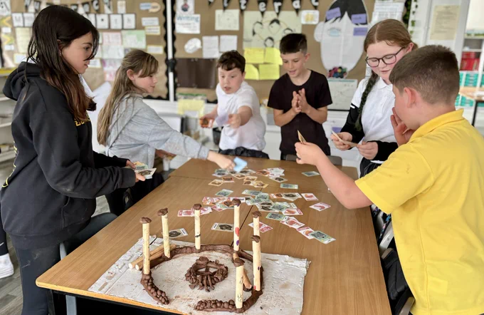 Photo of 6 animated pupils playing a card game, standing around a classroom table. Lots of cards on the table with pictures of animals. At the front of the table is a model of a timber circle.