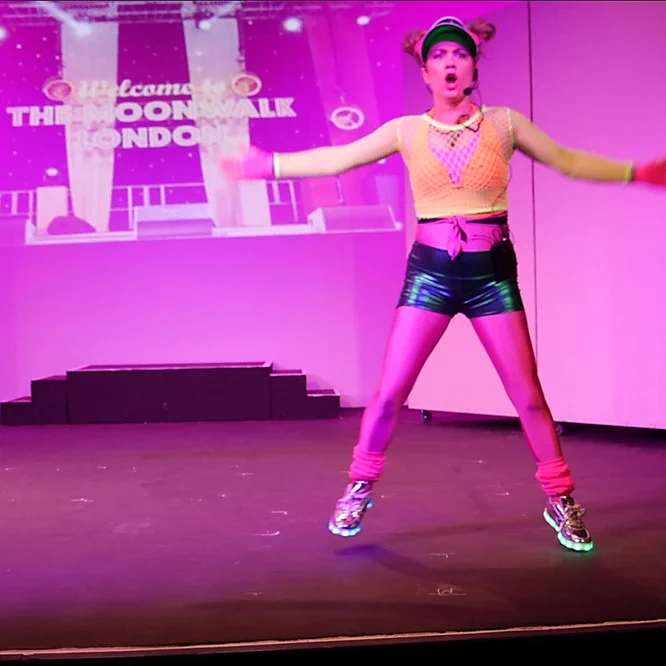 A member of the cast of Cheshire Cats, playing an aerobics instructor. She is jumping off the floor, arms in the air.
