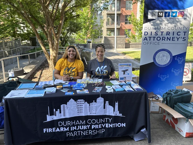 A photo of two women sitting at a table with gun locks and gun safety information. The tablecloth says “Durham county firearm injury prevention partnership.” A sign next to the table says “Durham county district attorney’s office”