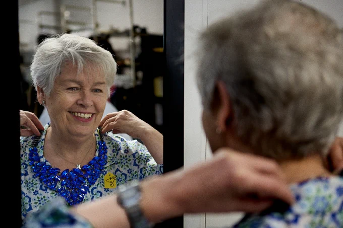 Maureen holding up a blue, jewelled necklace to herself in the mirror.