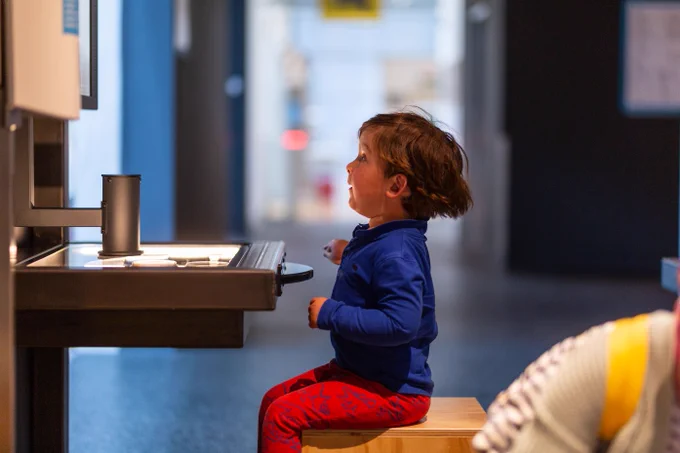 A young child in a blue shirt and red trousers sits on a stool in front of a table top exhibit at We The Curious.