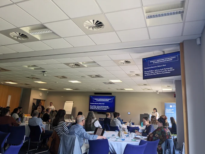 Connie at the speakers podium with delegates at tables facing the front with a screen with the sessions title improving inclusivity in marine decision making