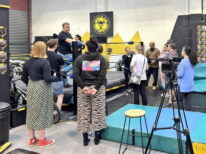 In a room with white walls, a group of people stand round a wrestling ring. There are 3 people in the ring, 2 fight and 1 talks to the group.