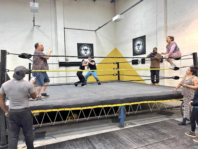 A wrestling ring in a room with white walls. In the ring are two people fighting, one person signing, one person sitting on the ropes, and one person filming with a camera. Two people watch from the floor.