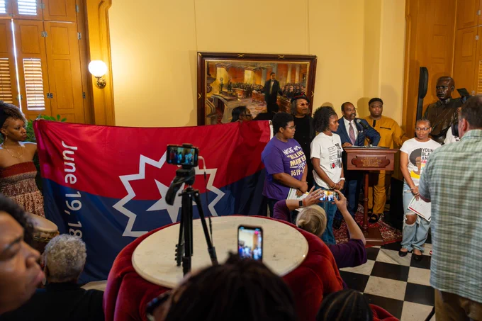 This is a photo of a group of people holding up a Juneteenth flag next to Senator Hearcel Craig speaking at a podium in the Ohio Statehouse.