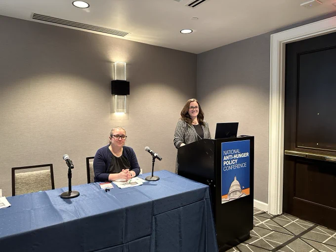 Two women presenting in conference room. One is standing at a podium and the other at a table. 