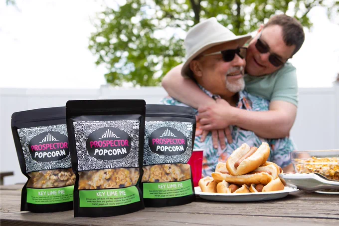 A son hugs his dad while they enjoy a picnic outside including hotdogs and Key Lime Pie gourmet popcorn.