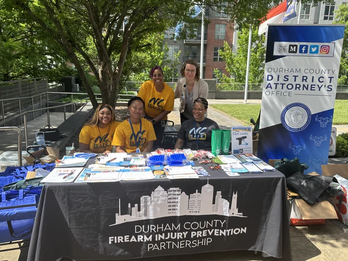 Five people, four of them wearing NC Safe t-shirts, sit at a table with gun locks and information. The tablecloth says "durham county firearm injury prevention partnership." A sign next to the table says "durham county district attorney's office."