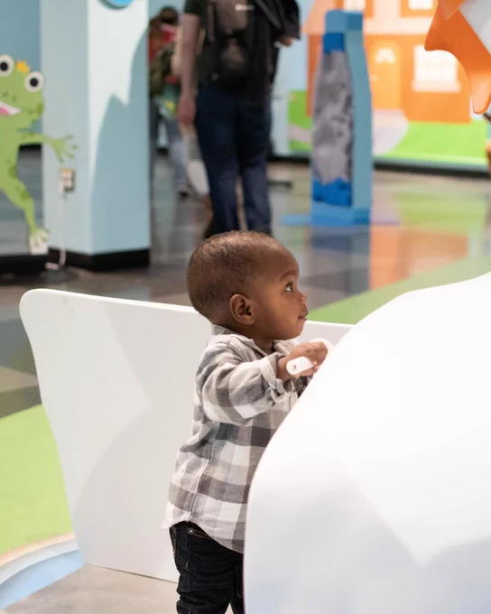 A young child, wearing a plaid shirt and dark jeans, is standing and holds onto an exhibit fixture in a brightly colored museum. The child is looking intently at something in front of them. The background features playful and colorful designs, including a large frog illustration on the wall and various other exhibits, with adults and children visible in the distance.
