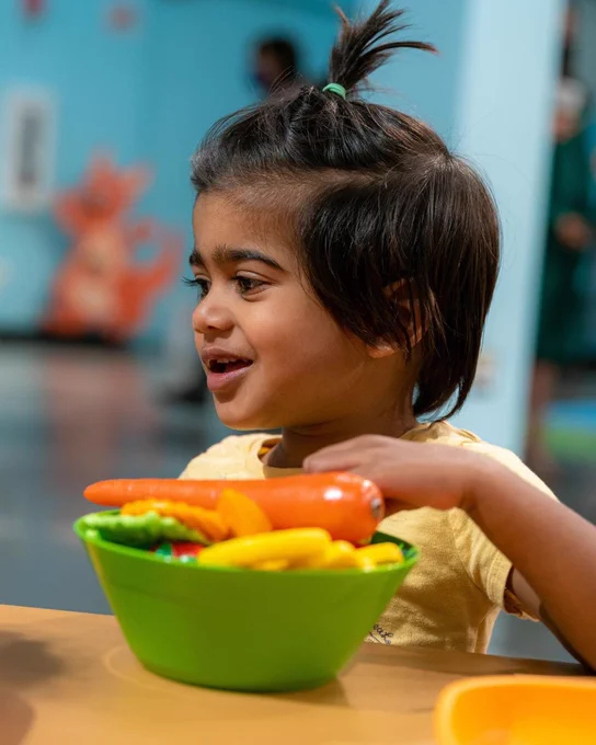 A young child with short dark hair tied in a small ponytail is sitting at a table in a museum. The child is smiling and looking to the side, engaging with a bowl of colorful toy vegetables. The background is brightly colored, with playful illustrations and other exhibits visible, along with a few blurred figures.