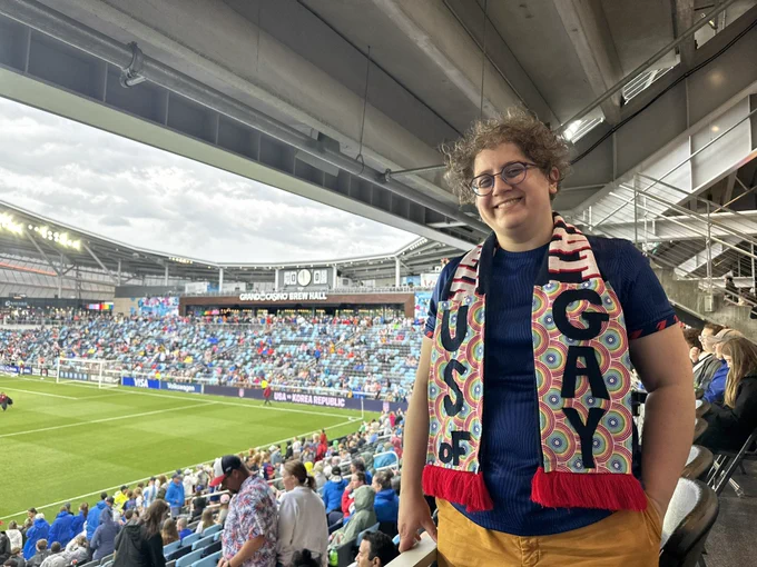 A white woman standing in the stands of a soccer stadium wearing a scarf that says “US of GAY”