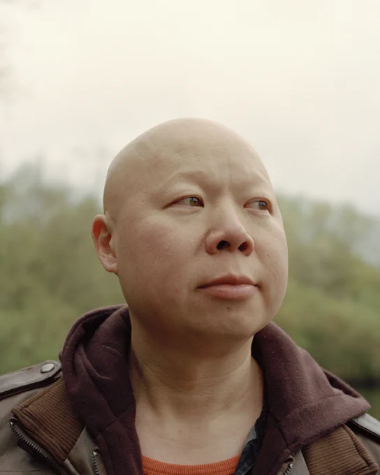 Raymond, a 30-40s year old man who has alopecia, wearing a brown jacket outdoors.