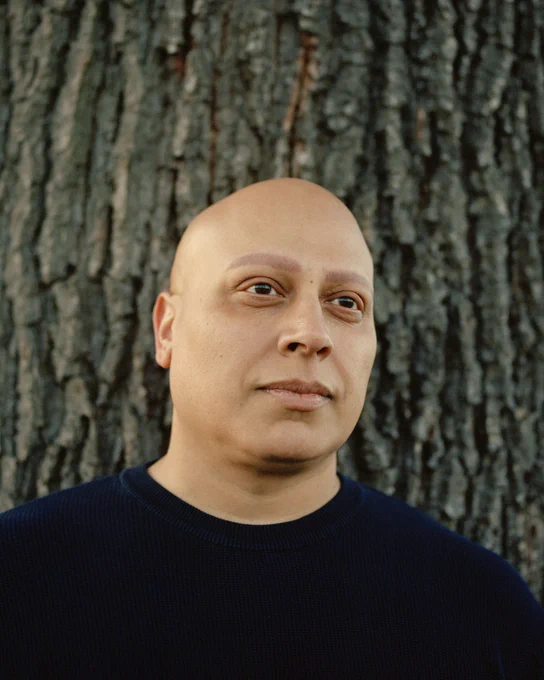 Bhavin, a man in his 40s who has alopecia, wearing a black jumper and standing in front of a tree.