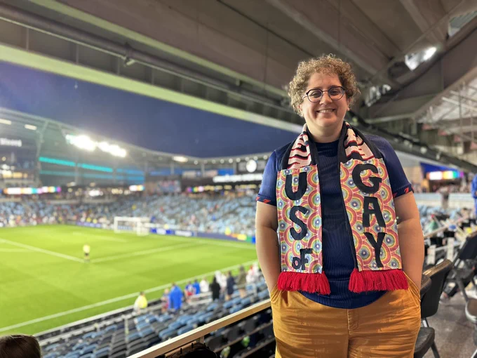 A white woman with curly brown hair standing in a soccer stadium with a soccer scarf that says “US of GAY.”