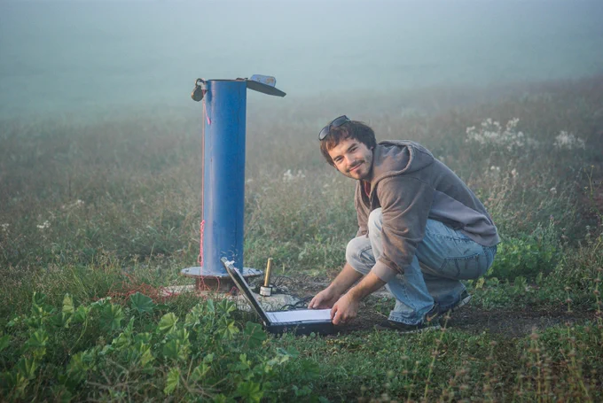 A man wearing jeans and a grey hoodie is crouched down working on a laptop next to a blue water pump in a misty, grassy field.