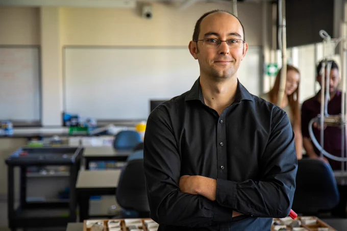 A man with glasses, standing confidently with folded arms, wearing a black shirt, in a classroom setting. There are students and laboratory equipment blurred in the background, suggesting an educational environment.