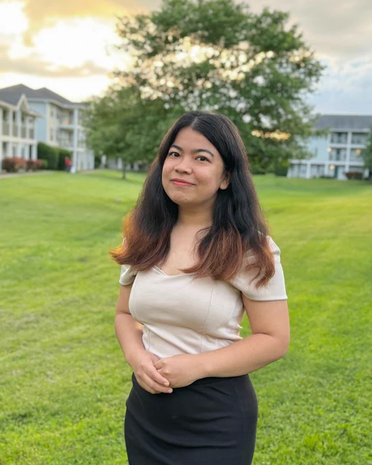 Aliching Marma stands in a court yard smiling, with a large tree and the clouds slightly orange, as if the sun is setting, behind her.