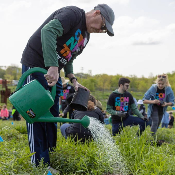 An individual waters plants using a green watering can, while other participants are engaged in gardening activities in the background.