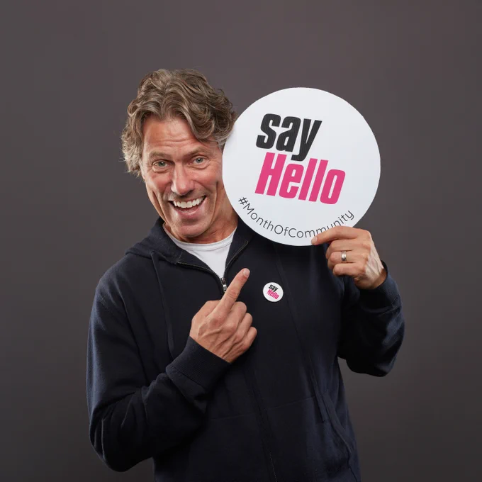 Comedian John Bishop holding and pointing to a sign saying say Hello #MonthOfCommunity