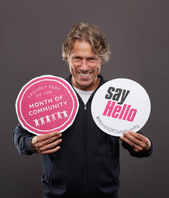 Comedian John Bishop holding 2 signs. One says Proudly part of the month of community. The other says Say Hello #MonthOfCommunity