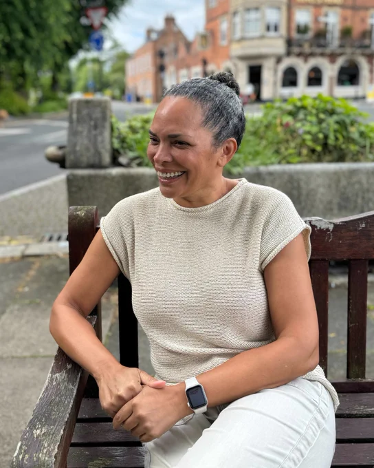 Nat smiling brightly, sitting on a bench, and looking into the distance.