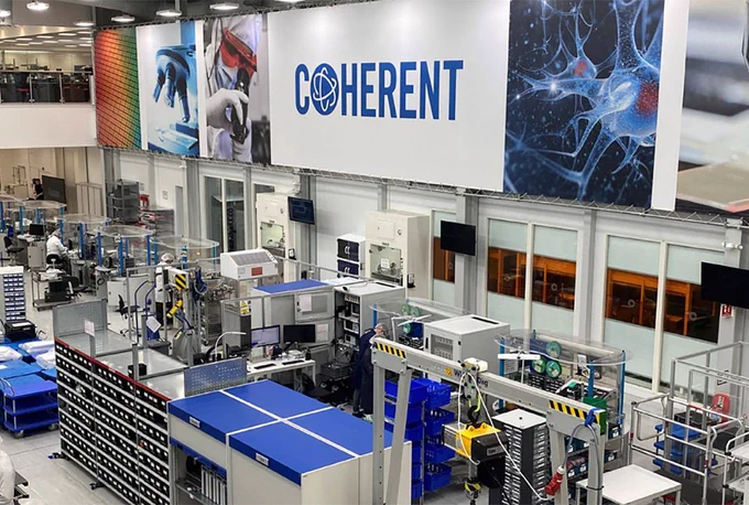 A manufacturing facility with various machinery and workstations is shown. Large "Coherent" signage hangs above, depicting scientific imagery.