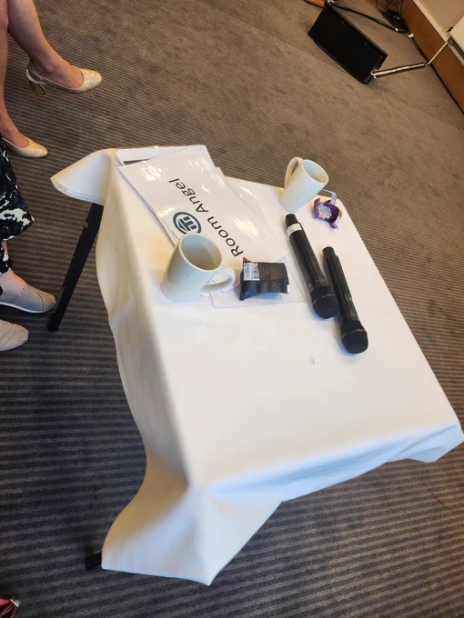 A table with a white tablecloth, two mics, a couple of ups of coffee and empty cookie packs, plus the "Room Angel" sign