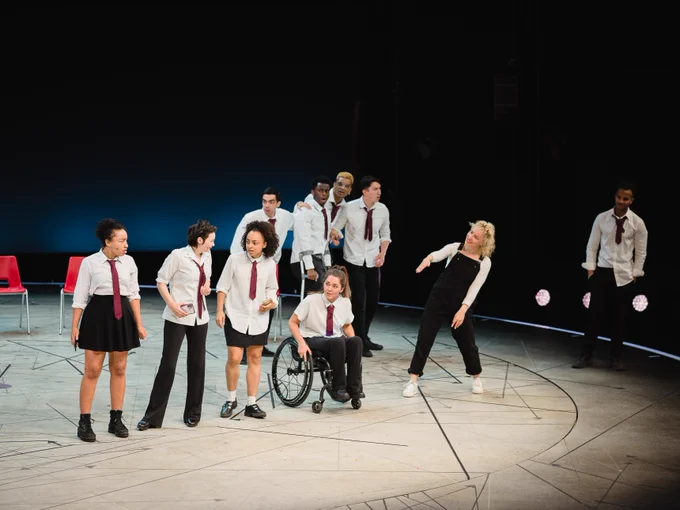 A group of performers on stage in a theatrical setting, with several wearing white shirts and dark trousers or skirts, and one performer in a wheelchair. One individual is dramatically leaning forward in a dynamic pose. The setting includes a simple background with scattered chairs and soft lighting emphasizing the central action.