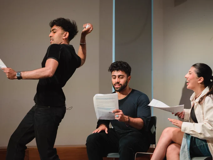 Three actors enthusiastically rehearsing a scene on stage, with one of them dramatically gesturing while holding scripts.