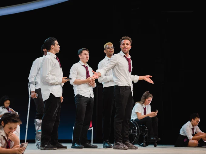 A group of five individuals in white shirts and dark pants, each wearing a maroon tie, are performing on stage. One person at the front is gesturing outward with arms extended, while the others stand close by with some holding hands. The setting includes other performers seated in the background, engaging in various activities.