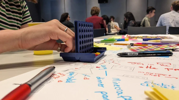 A photo of a person's hands playing mini connect 4 on top of a table covered in paper with written notes on game design with stationary, stickers and post-it notes also on the table. 