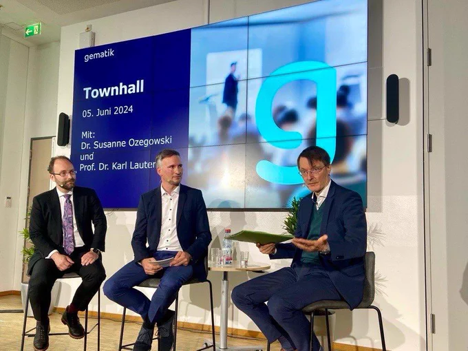 Bundesgesundheitsminister Karl Lauterbach und zwei Mitarbeiter der Gematik sitzen bei einem internen Townhall Meeting zusammen und diskutieren gemeinsam zum Thema Digitalisierung. Im Hintergrund ist ein Bildschirm mit der Aufschrift „Townhall - 5.6.2024, mit Dr. Susanne Ozegowski und Prof. Karl Lauterbach“ zu sehen.