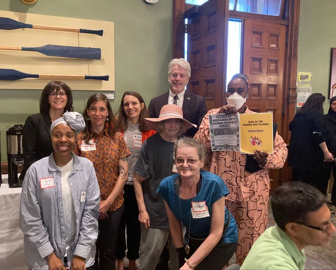 TNG Community Services representatives gather at the Disability Without Poverty's "Better the Benefit" campaign kickoff, along with Ontario MPPs Teresa Armstrong, Chris Glover and Jill Andrew. Jill holds up small posters promoting the event and campaign.