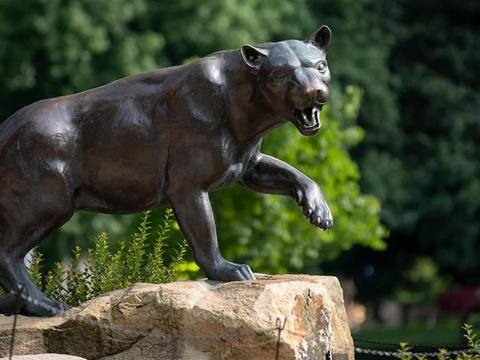 A bronze statue of a panther in a dynamic pose, positioned on a rock, set against a blurred background of green foliage.