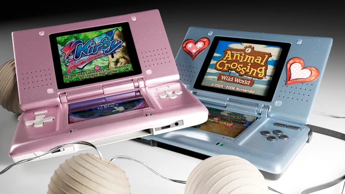 Render de dos modelos 3D de Nintendo DS antiguas, una rosa con un juego de Kirby en la pantalla y una Azul con pegatinas rojas de corazón y el juego de Animal Crossing Wild World en la pantalla. Rodeándolas hay una guirlanda de lucecitas también modelada en 3D.