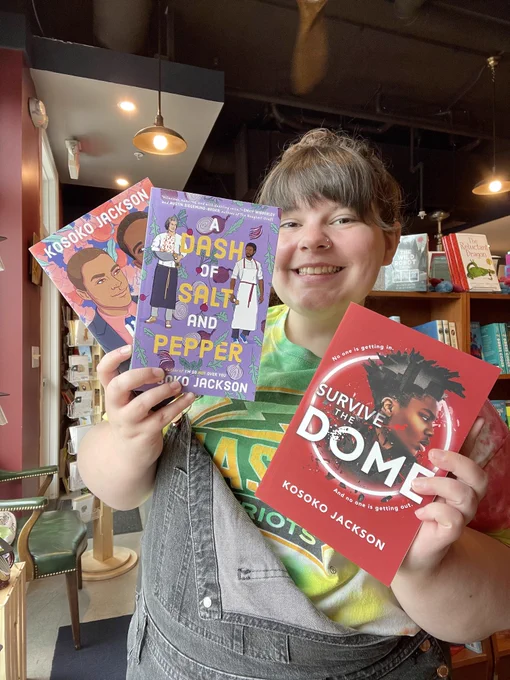 Bookseller Mallory holding up three books. From left to right they are I'm So (Not) Over You, A Dash of Salt and Pepper, and Survive the Dome, all by author Kosoko Jackson.