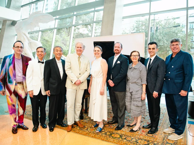 Group of nine people standing in an indoor setting with modern architectural elements, posing for a photograph beside a large art piece featuring abstract shapes. 
