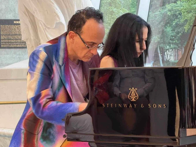 Two individuals are seated at a grand piano labeled "Steinway & Sons." The person on the left is dressed in a colorful jacket, while the person on the right, with long dark hair, appears focused on playing the piano. 