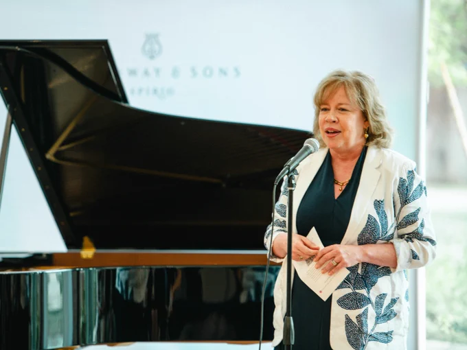 Person speaking at a podium with a grand piano. They hold a piece of paper and wear a white jacket with a floral design.