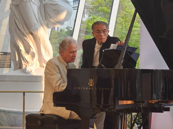 Person playing a Steinway & Sons piano at a public event with another person standing nearby, both in formal attire.