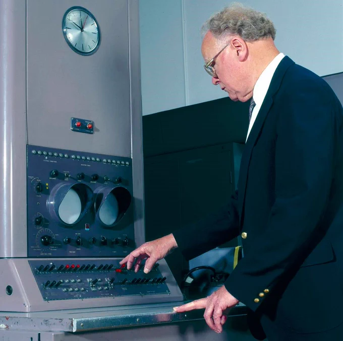 Photo of a white man in a dark suit with short curly hair pressing a button on the console of a Ferranti Pegasus computer in 1956 thanks to Science Museum Group Collection Online. (CC BY-NC-SA 4.0)
