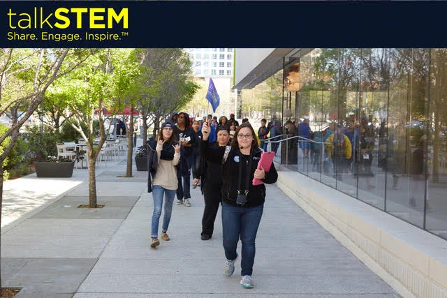 talkSTEM: Share. Engage. Inspire. Photo shows a group of people walking through an urban environment of mostly concrete and glass with a row of young trees alonside. The group's leader is wearing an ear microphone and holding a small blue flag high enough for the group to see and follow.