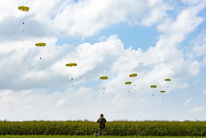 Paratroopers in the sky, with a serviceperson looking at them from the ground.