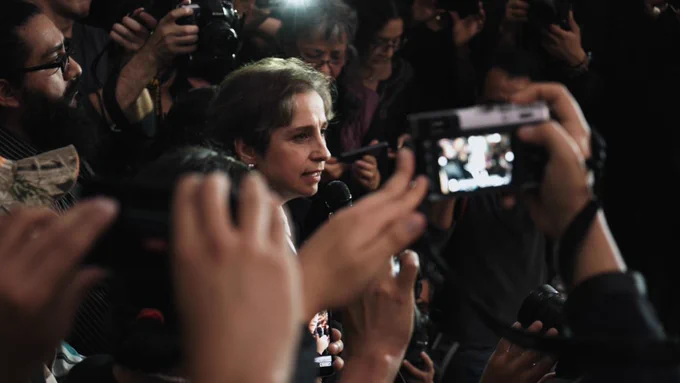 Carmen Aristegui speaks into a microphone in a thick crowd of people with cameras