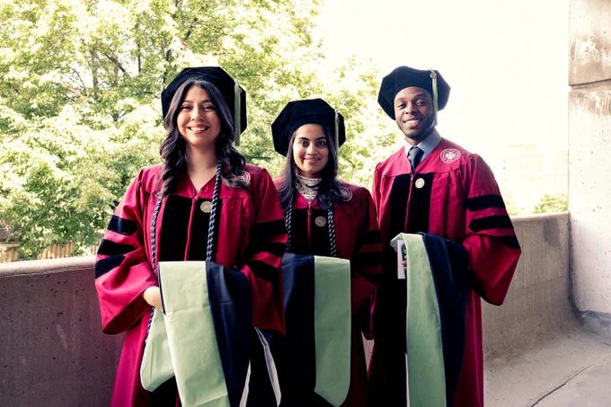 Three RFU graduates dressed in full regalia. They are standing next to each other in a parking garage. Behind them is an open space that reveals green trees and sunlight. 