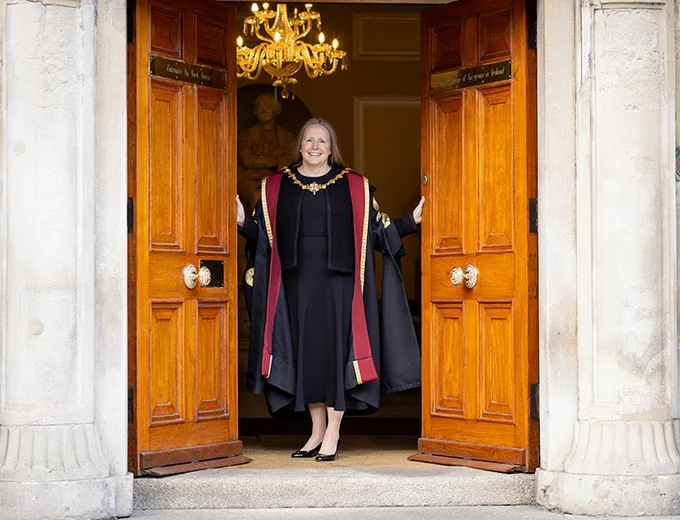 A photo of a person throwing open old wooden entrance doors. Visible behind her is an ornate hallway with a chandelier. She is wearing a cermonial gown and smiling broadly. 