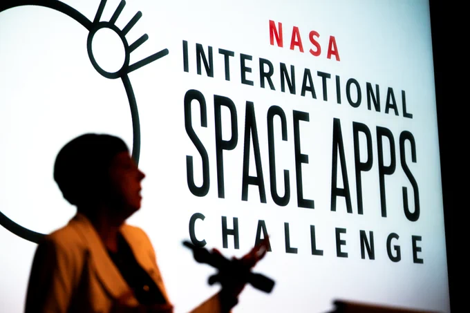 A large white screen reads "NASA International Space Apps Challenge", with a minimalist, circular logo to its left. A woman in a light-colored blazer, out of focus, stands in front of the screen on the far left of the photo. Credit: NASA/Keegan Barber