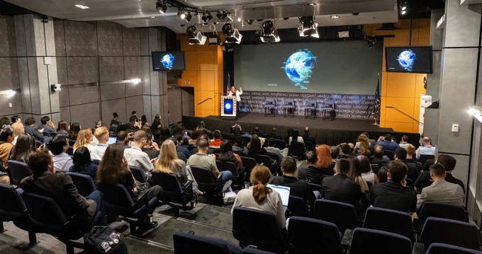 A crowd sits in a mid-sized auditorium, with dark seats and gray walls. At the front, a woman stands behind a podium with her left arm raised; on the screen behind her is a stylized image of Earth. Credit: NASA/Keegan Barber